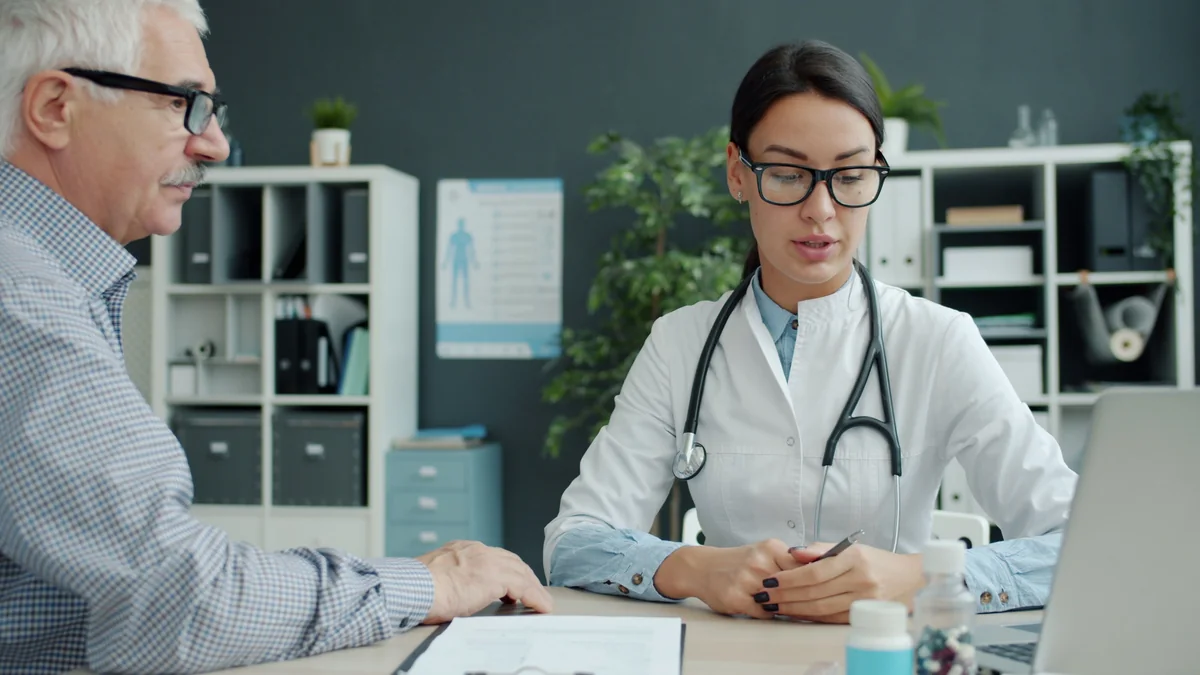 Older man in conversation with a clinician in a white coat across a desk in a consulting room