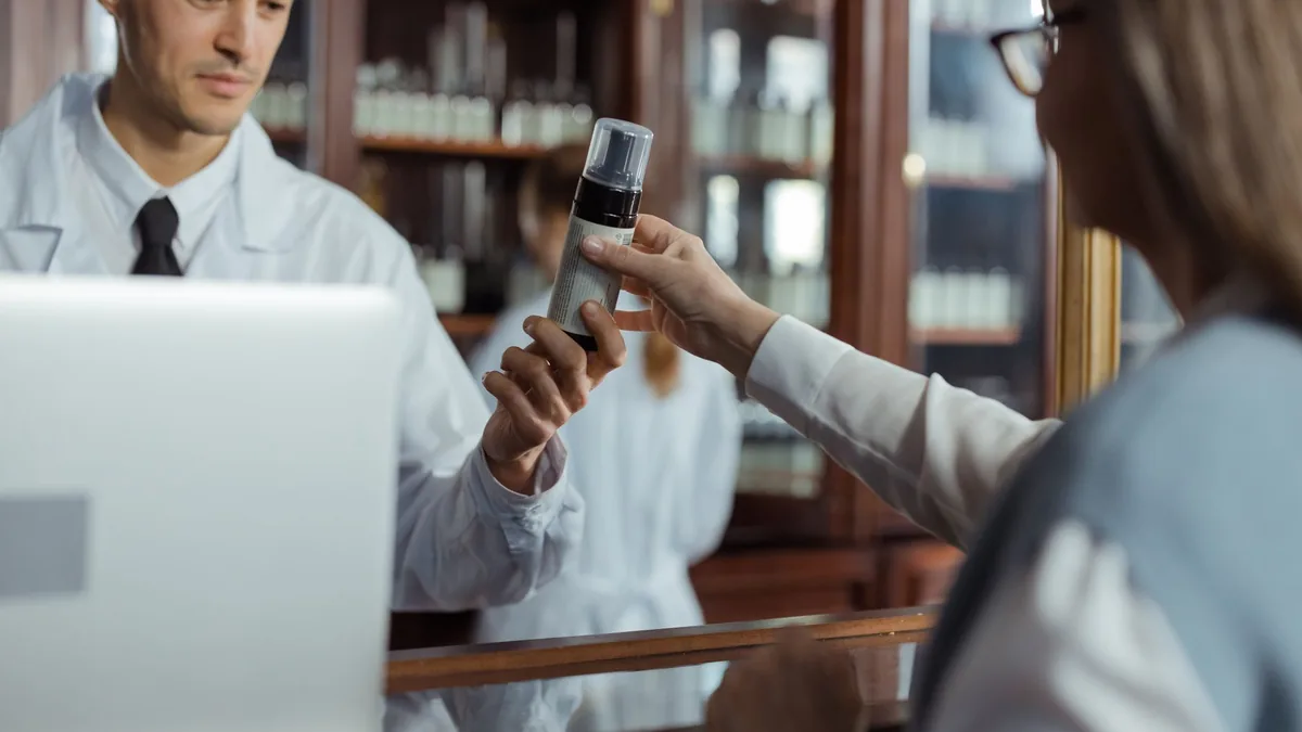 A pharmacist in a white uniform hands medication to a customer across the counter.